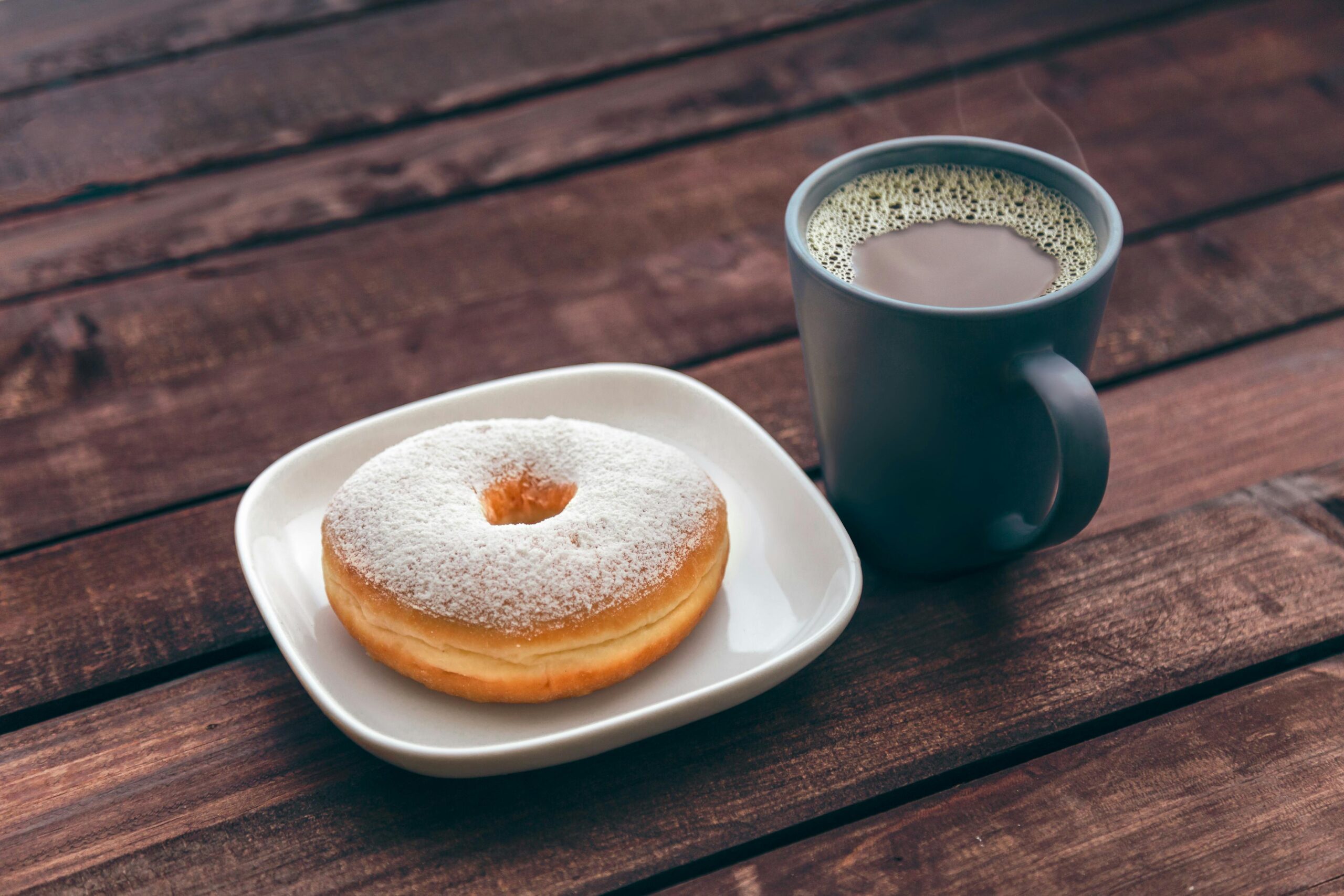 A steaming cup of coffee next to a powdered sugar donut on a wooden table.